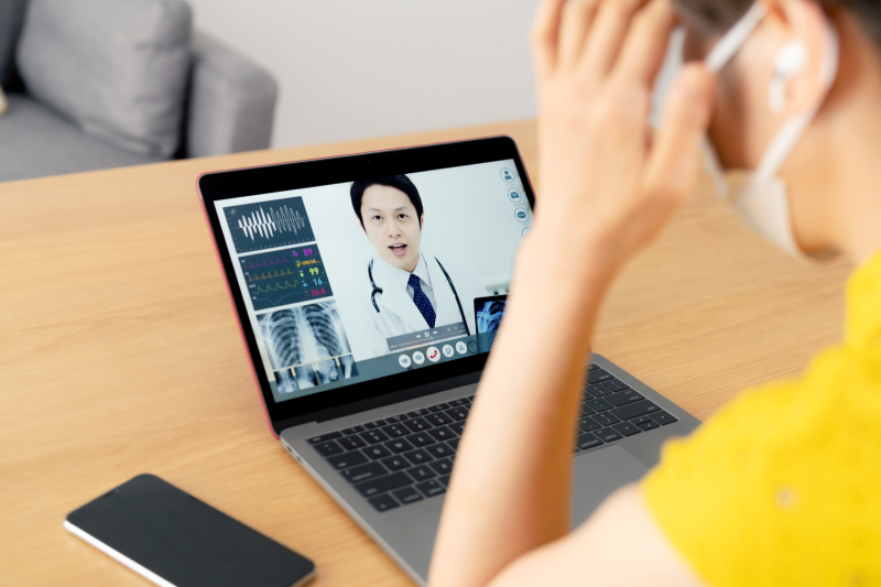 Side view of a woman having a video consultation with a male doctor through a laptop kept on a table and a mobile kept near it.