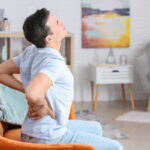 A young man sitting on a sofa holding his hands above the hip with a painful expression indicating a severe back pain.