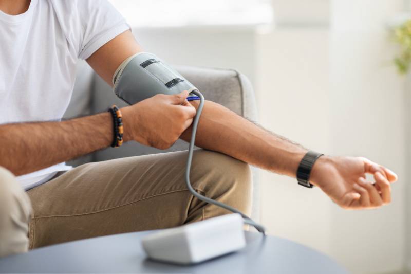 A man sitting on a chair with an automatic BP apparatus kept on a table before him and adjusting the cuff on his left hand.