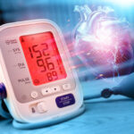 Image of an automatic blood pressure monitor showing readings along with a holographic image of a heart kept on a blue coloured table with the same colour themed background.