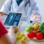 A nutritionist/female doctor using a tablet with graphical diagram of a human body along with a tray of vegetables,fruits and a measuring tape kept on the table explaining diet to a patient sitting before her.