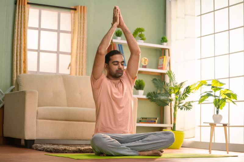 A man sitting on the floor of a room in a yoga posture with his hands held above his head in namaste pose depicting a healthcare routine.