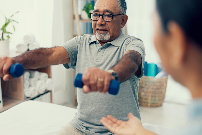 A physiotherapist guiding an elderly man doing exercise using dumbbell at nursing home/ rehab center.