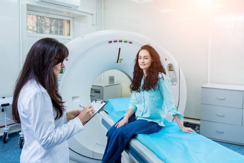 A female doctor holding a writing pad writes down the information given by the patient sitting on the CT table.