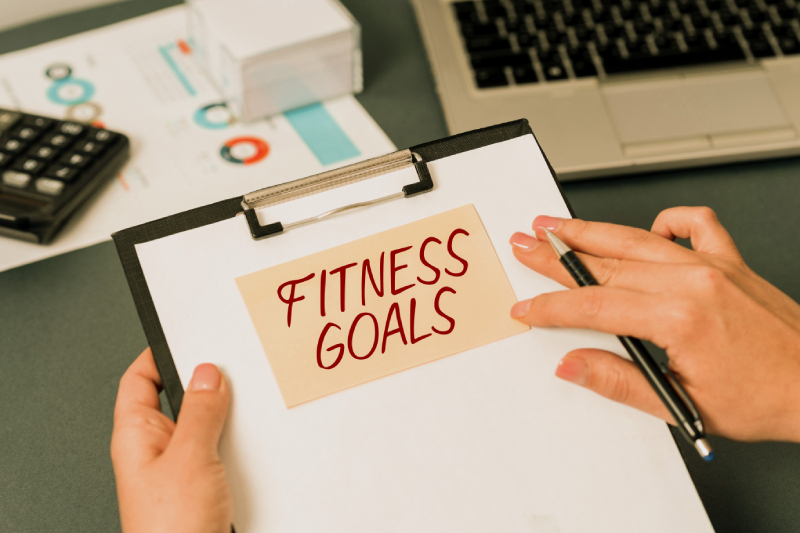 A persons hand holding a writing pad with the term FITNESS GOALS mentioned on it and other hand holding a pen.