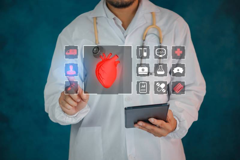 A male doctor holding a phone in his left hand touches the holographic image of a heart and related icons displayed before him.