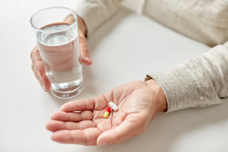 Close up image of a senior man sitting on a dining table holding pills and a water glass.