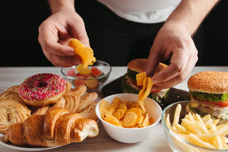 Junk foods kept on a table and a persons hands picking chips from the bowl signifying unhealthy food and overeating habits.