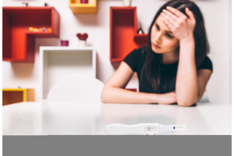 A depressed and sad looking woman sitting in a room holding her head and a pregnancy test kit showing negative result kept before her.