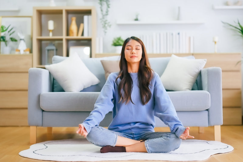 A young woman practicing/meditation at home sitting on living room floor in lotus position with closed eyes.