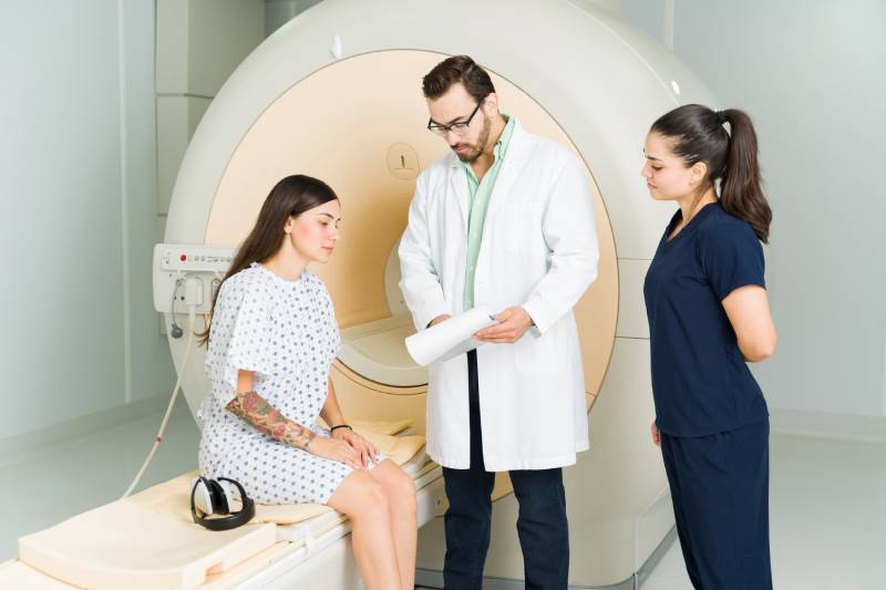 A male doctor along with a female technician shows a notepad and explains the MRI procedure to a female patient sitting on the MRI table.