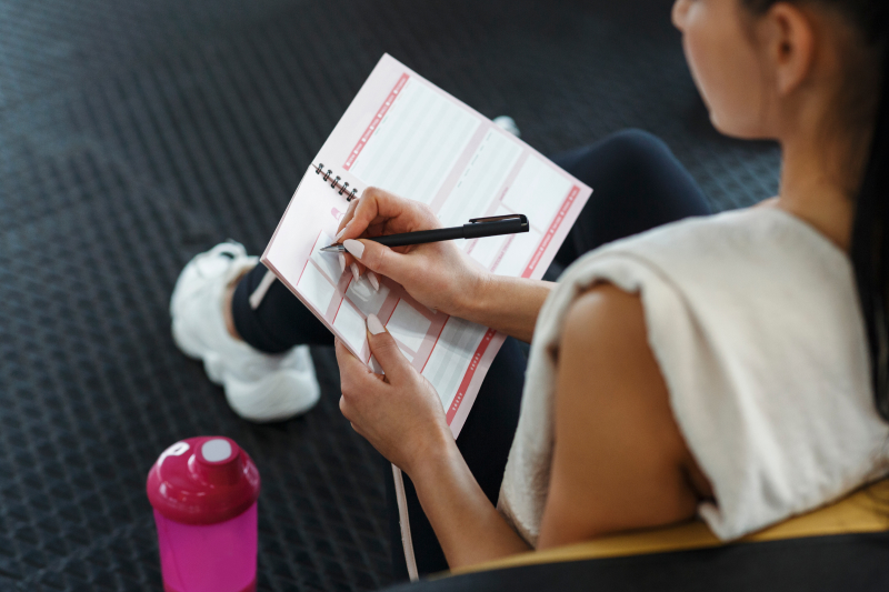 Rear view of a young woman sitting on a chair in a room making her workout schedule in a notebook and a water bottle kept near her.