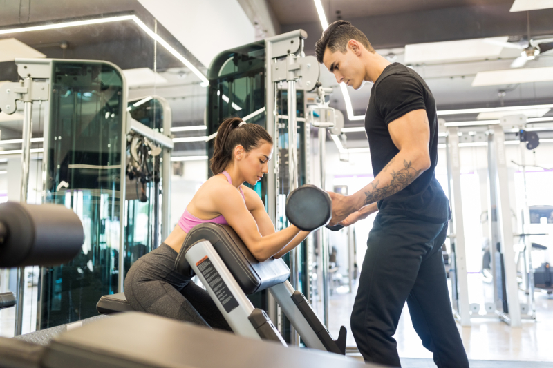 A male fitness instructor assisting his female client in lifting weight inside a modern gymnasium.