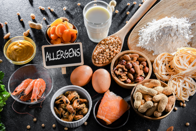 Top view of food items such as egg, milk, soya, nuts, fish, seafood, wheat flour, mustard, dried apricots and celery kept on a table with a tab displaying the term 