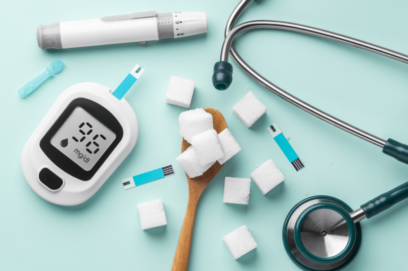 Top view of a pale blue coloured table with sugar cubes in spoon,blood glucose meter, lancet and stethoscope kept on it.