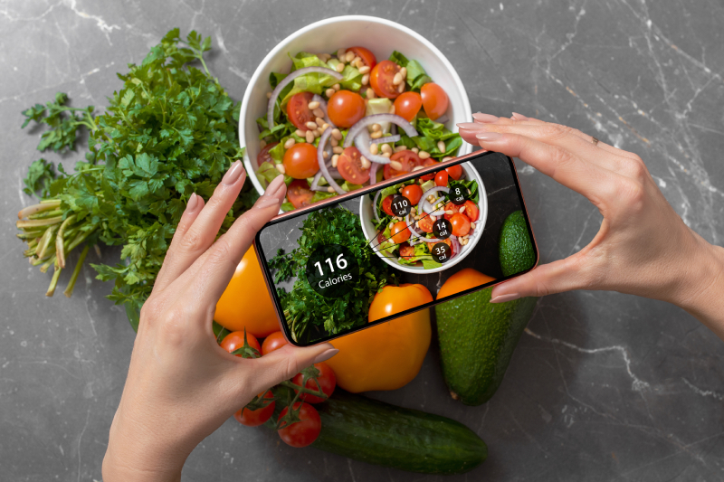 A female holding her smartphone above food items,using dieting app to track nutrition facts and calories in her food.