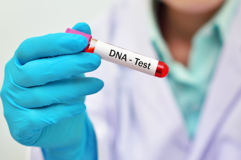 Close up image of a technician's hand wearing blue glove holding a blood sample tube with the abbrevation DNA mentioned on it.