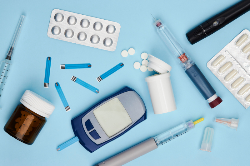 Top view of a glucometer, diabetic medicines, stripes and insulin syringe pen etc kept scattered on a pale blue background.