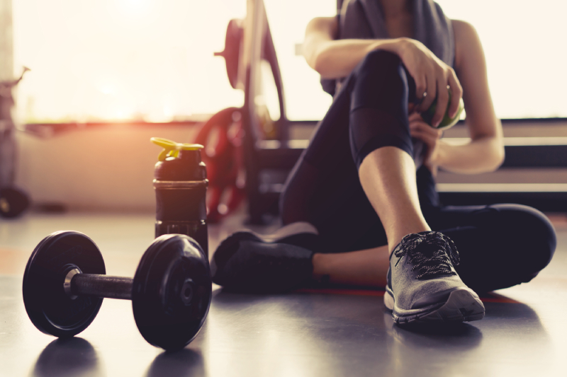 A woman sitting on the floor of a gym, holding an apple during a fitness break with a dumbbell and a water bottle placed near her.