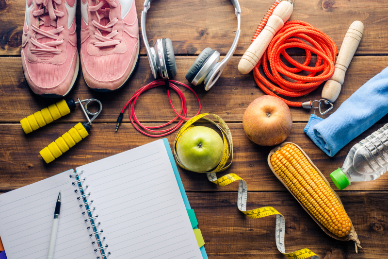 A pair of running shoes,skipping rope,headphones,apples,measuring tape,corn ,waterbottle and a towel kept along with a writing pad and a pen kept on a wooden table.