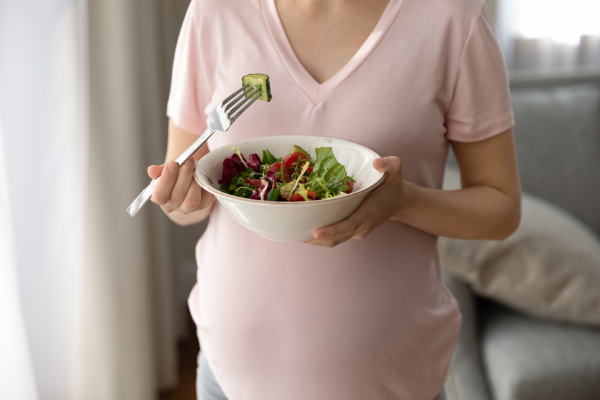 Close up view of an expecting woman holding a bowl of fresh vegetable salad and a fork with a piece of vegetable on it.