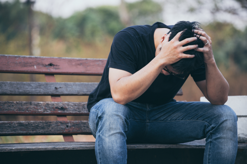 A blurred background with a depressed young man sitting on a wooden bench holding his head with his hands.