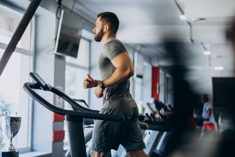 Side view of a young man using headphones running on treadmill facing the window, inside a gym.