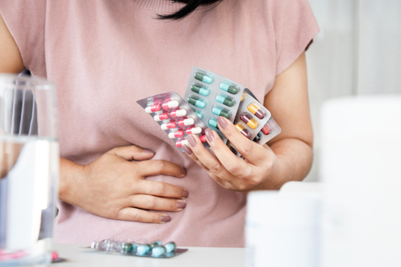 A close up image of a woman holding few capsule strips sitting on a bed/chair with an upset stomach due to side effect from overusing antibiotic medicines.