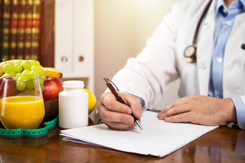 A nutritionist writing a diet plan on a chart kept on a wooden table and a bowl of fruits, fresh juice, medicine/supplements and a measuring tape kept near the chart.