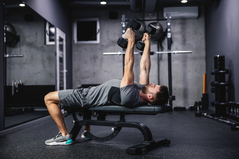 View of a gym with a young man lying on a dumbbell bench, lifting dumbbells in each hand.