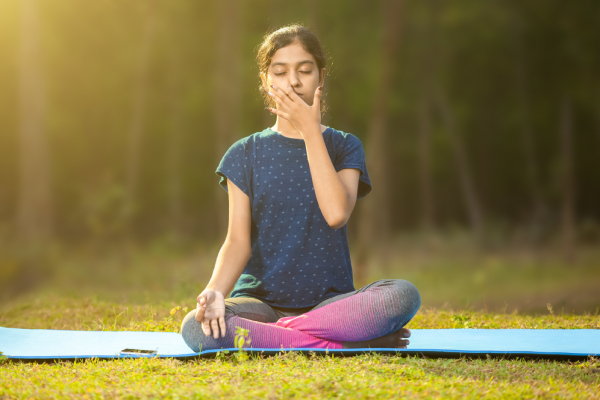 Blurred background view of a park/ground with a young woman doing pranayama sitting on a yoga mat.