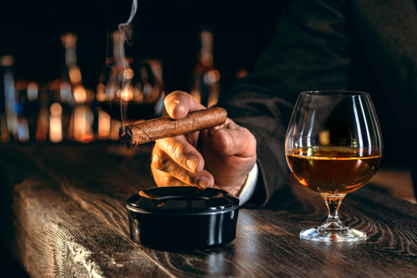 Image of a man's hand holding a cigar near an ash tray and a glass of alcohol kept on a wooden table in front of him , and blurred images of alcohol bottles displayed on a dark background beside him.