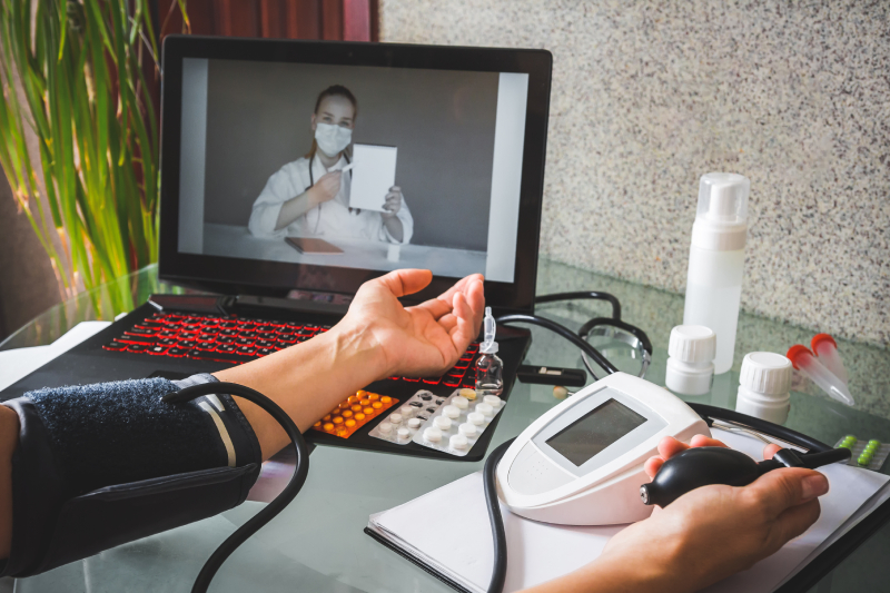 Image of a female patient's hands with the digital BP monitor measuring the readings meanwhile the doctor conducts a remote consultation and provides online medical assistance through the laptop in front of her.