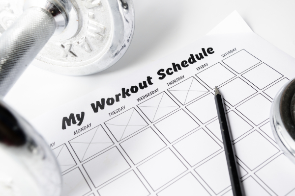 Image of a white sheet with day wise workout schedule placed on a table with a pencil and dumbbell kept on it.