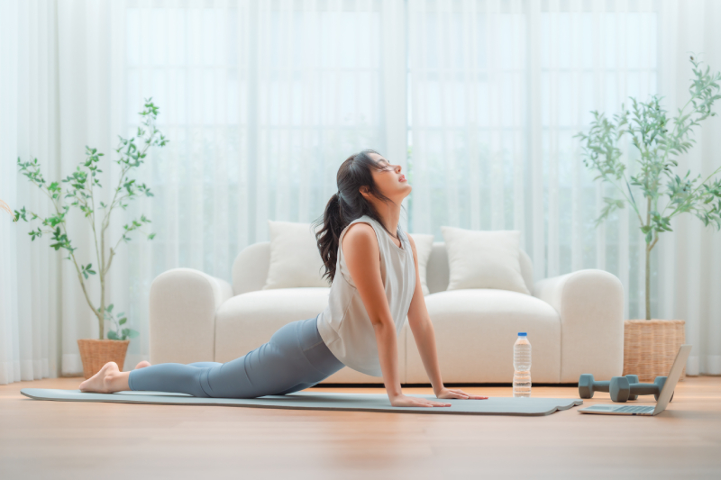 Image of a white themed living room with a woman/teenager lying on a yoga mat doing a stretching pose looking at a laptop and a pair of dumbbells and a water bottle kept near her.