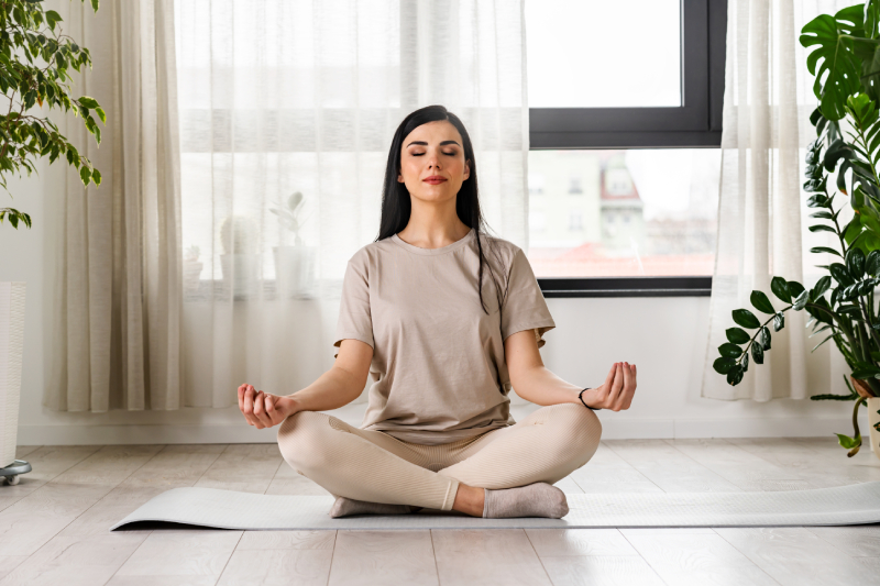 Indoor view of a white themed room with a young woman doing meditation, sitting on a yoga mat near a big window.