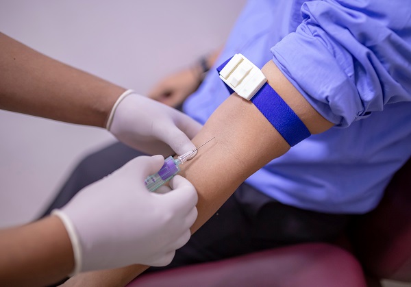 Image of a male patient sitting on a chair inside a laboratory and a technician withdrawing blood sample from his hand.