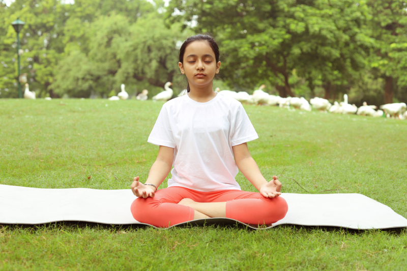 Front view of a child sitting on a yoga mat in a greenery filled park doing yoga.