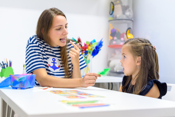 A female preschooler kid practicing correct pronunciation with the help of a female speech therapist using different coloured objects kept on the table.