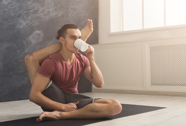 A young sitting on mat in a flexible yoga pose sipping water/tea.