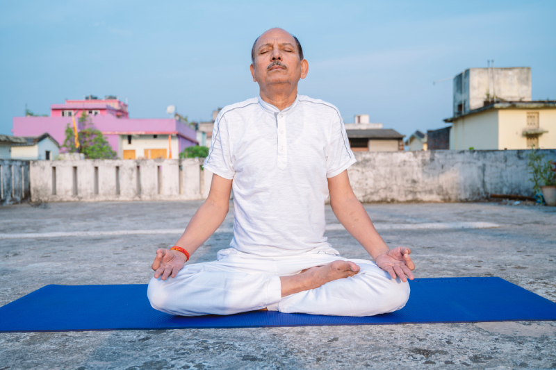 An elderly person in white dress, sitting in yoga pose on a terrace.