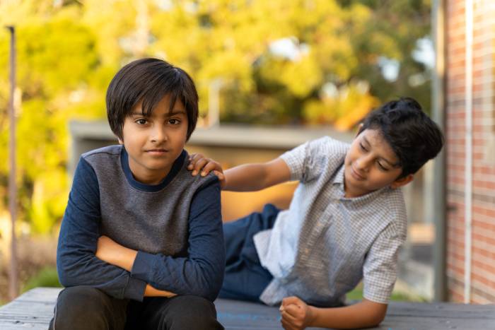 A boy sits with his hands together, and his friend behind him rests one hand on the ground and the other on his shoulder in an outdoor setting.