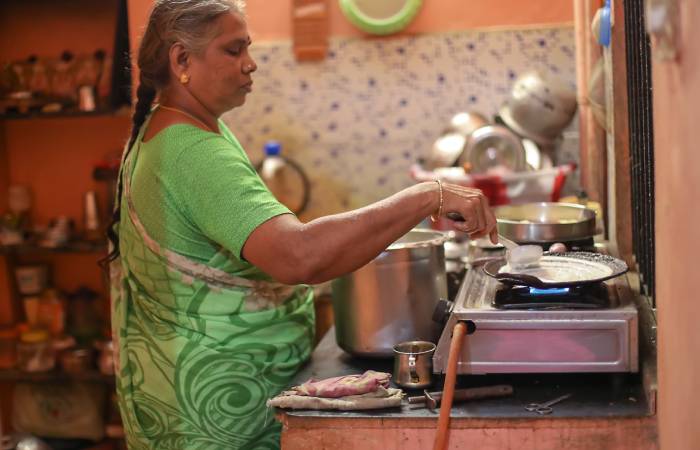 An elderly Indian woman making dosa inside an ordinary kitchen.