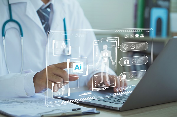 A doctor using a laptop and a writing pad kept on a table before him and his right hand pointing at a hologram of AI related concepts in healthcare displayed above the keyboard.