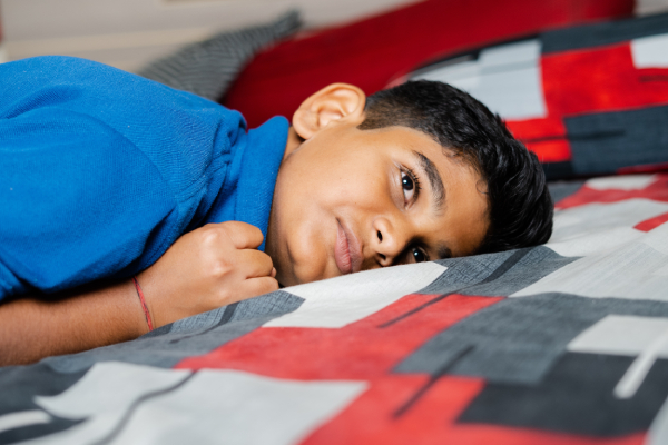 A boy lies on the bed in an upside-down position, facing one side and looking sad.