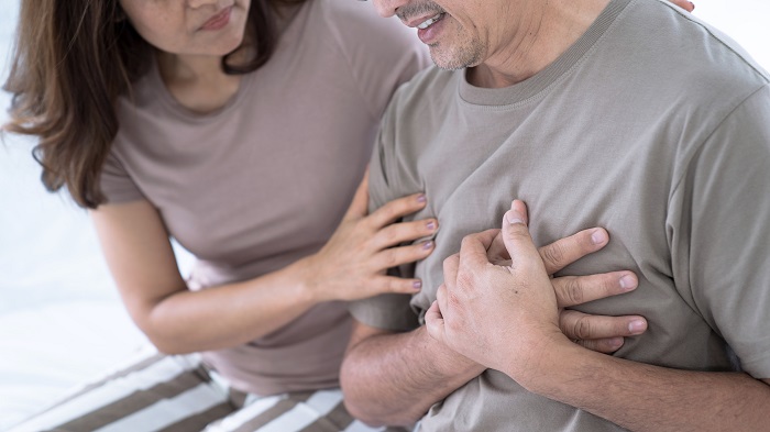 A middle aged man sitting with his hands holding the left side of his chest in pain and a woman attending on him.