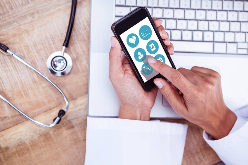 A doctor is using a medical app on their mobile phone in front of a laptop on the table.