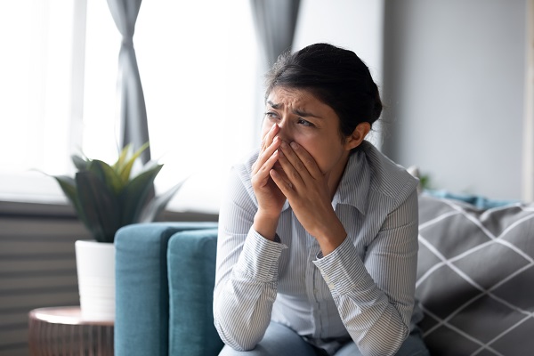 Image of a living room with a woman sitting on a sofa, with her hands covering her lower face in a sad/depressed condition.