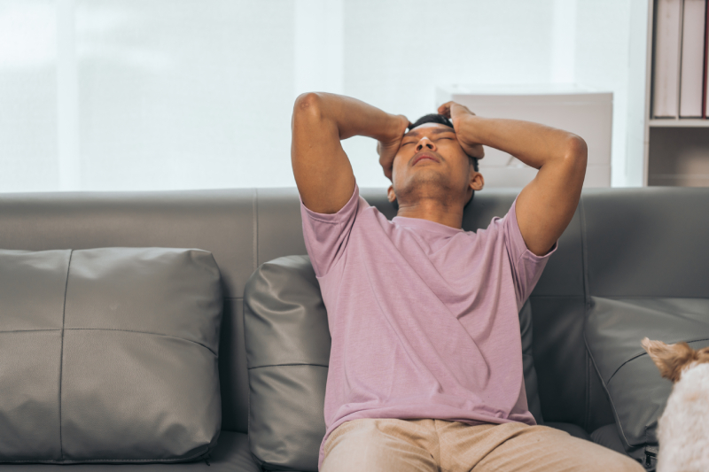 A man sits on the sofa with his eyes closed, holding his head with both hands as he thinks.