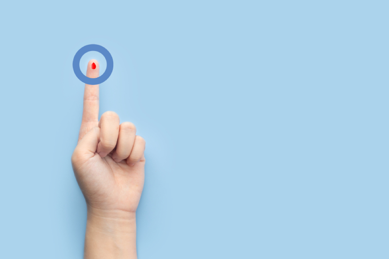 Image of a persons hand on a blue background and the index finger with a blood drop and blue circle around it representing the universal symbol for diabetes.